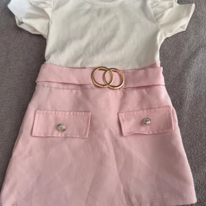 Söt kortklänning med bälte och fickor - Supersöt kortklänning med vit ribbad överdel och puffärmar, samt ljusrosa kjol med dekorativa fickor och guldigt bälte framtill. Kjolen är i mjuk polyesterblandning och klänningen har en riktigt gullig och trendig look.