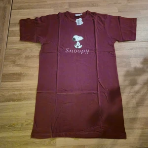 Vinröd Snoopy t-shirt i bomull - Säljer en vinröd t-shirt med tryck av Snoopy på bröstet. Kan nog också användas som nattlinne eller pyjamasöverdel beroende på storlek. T-shirten är gjord i 100% bomull och har en klassisk rund hals samt korta ärmar. Passformen är normal och materialet är mjukt och skönt mot huden. Perfekt för dig som gillar Peanuts och vill ha en chill stil. Varan är helt oanvänd med etiketten kvar.
