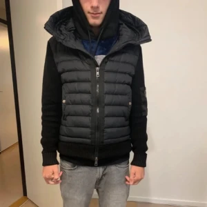 Svart pufferjacka från Moncler - Svart pufferjacka från Moncler med stickade ärmar och quiltade detaljer. Jackan har huva, dragkedja framtill och en extra ficka med dragkedja på ärmen. Klassisk Moncler-logga på vänster ärm. Perfekt för kyliga dagar.