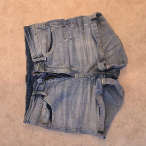 Ljusblå jeansshorts med dragkedja - Säljer ett par ljusblå jeansshorts med klassisk femficksdesign och dragkedja framtill. Shortsen har en avslappnad passform och slitna detaljer på framsidan för en cool look. Perfekta för varma dagar och enkel att matcha med t-shirt eller linne.