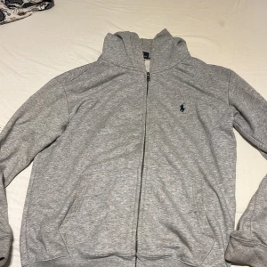 Grå hoodie från Polo Ralph Lauren - Grå hoodie från Polo Ralph Lauren med dragkedja framtill och klassisk blå broderad logga på bröstet. Tröjan har huva, långa ärmar och två fickor framtill. Tillverkad i mjuk bomull för en chill och bekväm stil.