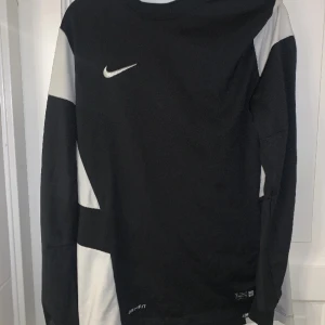 Nike Dri-Fit svartvit långärmad tröja - Svart och vit långärmad tröja från Nike med broderad swoosh på bröstet och 'Authentic Nike Team'-märke vid nederkanten. Tillverkad i Dri-Fit polyester som andas, perfekt för träning eller sport. Snygg sportig design med kontrastpaneler på ärmarna.