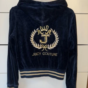 Vintage Juicy Couture marinblå velour hoodie - Juicy Couture hoodie i marinblå velour med guldfärgade broderier och strassdetaljer. Dragkedjan har en stor J-berlock och tröjan har huva samt ribbade muddar med guldiga ränder. Klassisk logga på ryggen och mindre tryck i fram. Några paljetter som lossnat men annars fint skick.