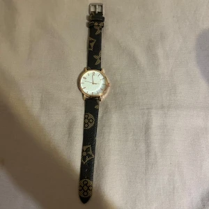 Svart klocka med mönstrat läderband - Stilren klocka med vitt ur och svart läderband dekorerat med guldiga mönster. Urtavlan har romerska siffror och boetten är i guldton. Perfekt accessoar för att lyfta din outfit.