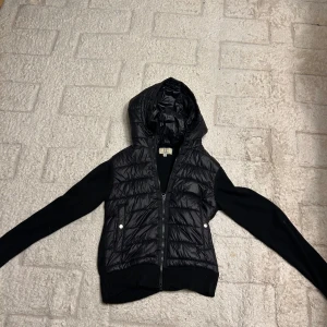 Svart pufferjacka från Elegance Essentials XS - Svart pufferjacka med stickade ärmar och huva från Elegance Essentials. Jackan har quiltad front i glansigt material, dragkedja och två fickor med tryckknappar. Perfekt för dig som gillar en clean och stilig look. Ny pris är 1200 men den år slutsåld. Den passar runt 165-170.