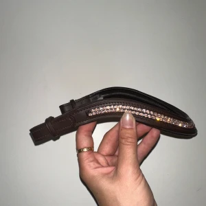 Brunt pannband med stenar. Full - Mörkbrunt pannband i skinn med glittriga stenar i rosé. Går att knäppa på superenkelt med kardborrefästen i sidorna. Helt oanvänt! Storlek Full. 