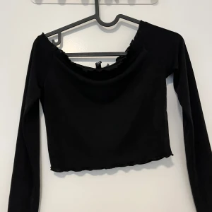 Svart croppad långärmad topp - Snygg svart croppad topp med långa ärmar och offshoulder-skärning. Toppen har en lätt vågig kant vid halsringningen och nederkanten, vilket ger en trendig look. Perfekt att styla med höga jeans eller kjol.