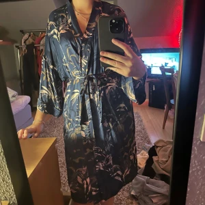 Mörkblå omlottklänning med blommor - Snygg mörkblå omlottklänning med blommigt mönster och knytband i midjan. Klänningen har vida trekvartsärmar och ett lyxigt, glansigt material som känns riktigt skönt mot huden. Perfekt för dig som vill ha en chic och avslappnad look.