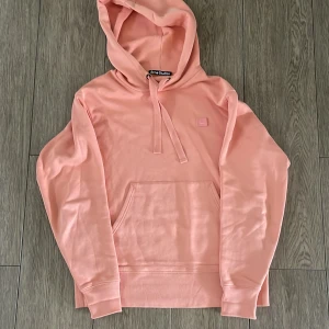 Rosa hoodie från Acne Studios - Säljer en snygg ljusrosa hoodie från Acne Studios i 100% bomull. Tröjan har huva med snörning, magficka och det klassiska Face-logotypen broderad på bröstet. Perfekt för dig som gillar stilrena och trendiga plagg.