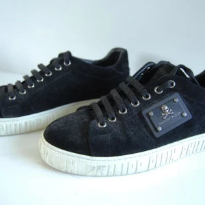 Philipp Plein svarta mockasneakers - Svarta sneakers från Philipp Plein i mocka med vita sulor där märkesnamnet är präglat runtom. Skorna har snörning med metallöljetter och en metallplatta med dödskalle på sidan. Perfekta för dig som gillar en edgy och lyxig stil. Ny pris 5500 mycket bra skick använda 5 gånger