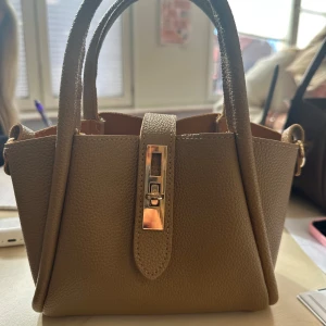 Beige handväska med gulddetalj - Snygg beige handväska i skinn med guldfärgad låsdetalj framtill. Väskan har två handtag och en stilren, boxig form som passar perfekt till din vardagslook. Enkel men elegant design som lyfter vilken outfit som helst. Säljer för 250kr! 