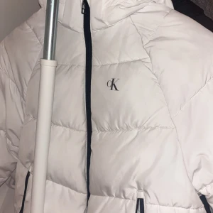 Vit pufferjacka från Calvin Klein - Snygg vit pufferjacka från Calvin Klein med svart dragkedja och huva. Jackan har CK-logga på bröstet och svarta detaljer vid fickorna. Perfekt för kalla dagar och ger en clean, modern look. Materialet är syntet och jackan är quiltad för extra värme.