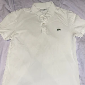 Vit pikétröja från Lacoste XS - Klassisk vit pikétröja från Lacoste i slim fit-modell. Tröjan har korta ärmar, krage med knappar och den ikoniska gröna krokodilloggan broderad på bröstet. Tillverkad i bomull för en skön och luftig känsla.