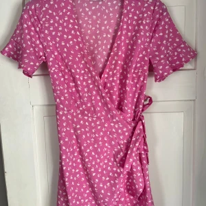 Rosa klänning  - Supersöt rosa omlottklänning med vitt blommigt mönster och knyt i midjan. Klänningen har korta ärmar med volangkant och v-ringad hals. Perfekt för varma dagar och ger en fräsch och somrig vibe. Använd få gånger. 