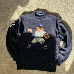 Polo Bear tröja från Ralph Lauren - Mörkblå stickad tröja från Polo Ralph Lauren med ikoniska Polo Bear i basebolloutfit på bröstet. Tröjan har rund hals, långa ärmar och ribbade muddar. Materialet är en lyxig mix av ull, kashmir och kamelhår. Perfekt för dig som vill sticka ut med sportig preppy-stil. Sparsamt använd.