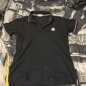 Svart Moncler piké t-shirt M - Svart piké t-shirt från Moncler i bomull med klassisk krage och korta ärmar. Röd, vit och blå rand på krage och ärmslut samt Moncler-logga på bröstet. Snygg och stilren modell som passar perfekt till jeans eller shorts.