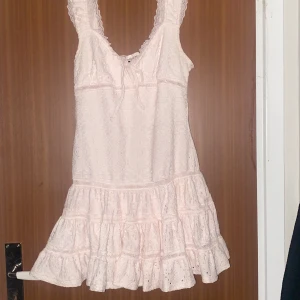 Ljusrosa spetsklänning från Gina Tricot - Supersöt ljusrosa kortklänning från Gina Tricot med breda axelband, volangkant nertill och v-ringning med knyt. Klänningen är i spets med broderade detaljer och har en romantisk vibe. Perfekt för sommarens alla tillfällen!