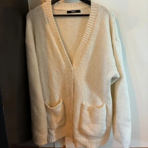 Beige stickad kofta från Bik Bok - Mysig beige stickad kofta från Bik Bok med djup v-ringning och två stora fickor framtill. Perfekt för lager-på-lager och har en avslappnad, loose passform med långa ärmar och bred ribbad kant.