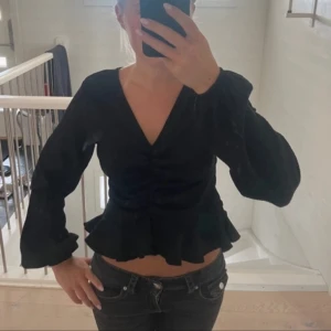Svart croppad blus med volang - Snygg svart blus med v-ringning, långa puffiga ärmar och volangkant nertill. Toppen är croppad och har rynkade detaljer framtill som ger en trendig look. Perfekt att styla med jeans eller kjol för en cool outfit.