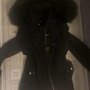 Jacka från zara - Snygg svart parkajacka med stor huva och fluffig fuskpäls. Jackan har coola spänndetaljer, dragkedja framtill och stora fickor med knappar. Perfekt för kalla dagar och ger en riktigt trendig look. Jackan är i bra skick