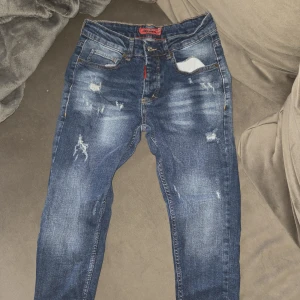 Dsquared2 blå slitna jeans - Snygga blå jeans från Dsquared2 med slitningar och distressed detaljer framtill. Klassisk femficksmodell med röda sömmar och Dsquared2-logga på insidan. Jeansen har en rak passform och är tillverkade i jeansmaterial.