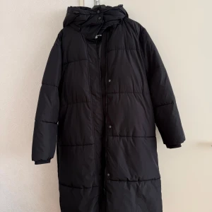 Svart lång pufferjacka med huva - Säljer en svart lång pufferjacka med huva och bälte i midjan. Jackan har quiltad design, dragkedja och knappar framtill. Perfekt för kalla dagar när du vill hålla dig varm och ändå se stilren ut. 