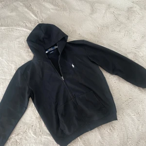 Svart hoodie från Polo Ralph Lauren - Svart hoodie från Polo Ralph Lauren med vit broderad logga på bröstet. Tröjan har huva, dragkedja framtill och två stora fickor. Tillverkad i mjuk bomull, perfekt för chill dagar. Custom fit-modell som sitter snyggt och bekvämt.