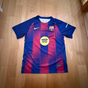 FC Barcelona Nike matchtröja M - Säljer en officiell FC Barcelona matchtröja från Nike i storlek M. Tröjan har klassiska blå och röda vertikala ränder, klubbmärke på bröstet, gul Nike-logga och sponsortryck från Spotify. Tillverkad i Dri-FIT-material som andas, perfekt för fotboll eller träning.