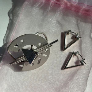 Geometriska örhängen och brosch i silver - Säljer ett set med två triangelformade örhängen och en stor, rund brosch med futuristisk design i silverfärgad metall. Broschen har inslag av cirklar, trianglar och raka linjer för en cool, modern vibe. Perfekt för dig som gillar unika accessoarer.