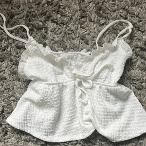 Vit linnetopp med volang och knyt - Supersöt vit linnetopp med smala axelband, volangkant upptill och knytning framtill. Toppen har en lätt rynkad struktur och är perfekt för varma dagar. Den är ärmlös och har en luftig, croppad passform som ger en fräsch och somrig vibe.