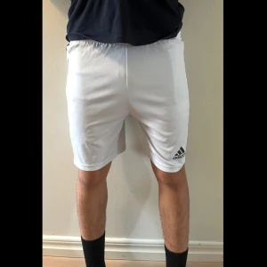 Vita Adidas shorts med svarta ränder - Säljer ett par vita Adidas shorts med klassiska svarta ränder längs sidorna och elastisk midja. Lätta och sköna i syntetmaterial, perfekta för träning eller fotboll. Adidas-logga framtill och AEROREADY-tryck bak. Storlek: 13-14 år (164)