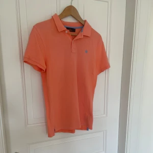 Orange pikétröja från Morris, stl M - Snygg orange pikétröja från Morris i storlek M. Klassisk krage, korta ärmar och broderad logga på bröstet. Tillverkad i bomull och har en fräsch, stilren look med blå detalj på insidan av kragen.