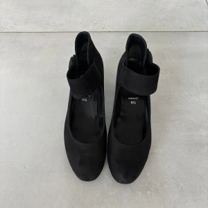 Svarta mocka pumps från Gabor - Snygga svarta pumps från Gabor i mocka med rund tå och låg klack. Skorna har en bred rem med kardborreknäppning över vristen och en dekorativ knapp på sidan. Perfekta för dig som gillar klassisk och stilren design med en twist.