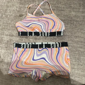 Färgglad bikini från DEX, L.A. Surf - Säljer en bikini från DEX x L.A. Surf med coolt vågigt mönster i lila, orange, rosa och beige. Toppen har korsade axelband och bred resår med logga. Underdelen är shortsmodell med samma resår. Tillverkad i återvunnen polyesterblandning.