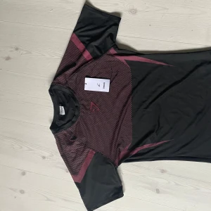 Svart och vinröd Gymshark t-shirt onyx v5 S - Snygg svart t-shirt från Gymshark i storlek S med vinröda detaljer och grafiskt mönster över bröst och rygg. Tillverkad i mjukt och lätt polyestermaterial, perfekt för träning. Klassisk rund hals och korta ärmar för en sportig vibe. Passar dig perfekt om du har M i T-shirts då onyx v5 är lite större i storleken