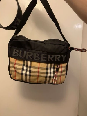 Burberry rutig axelväska med logga - Säljer en snygg axelväska från Burberry med klassiskt rutigt mönster i beige, svart, rött och vitt. Väskan har en bred justerbar axelrem med Burberry-logga och dragkedja upptill. Materialet är en mix av bomull och syntet, perfekt för att bära dina essentials.