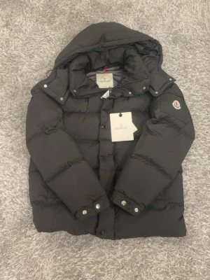 Moncler jacka - Svart pufferjacka från Moncler med huva, snygga detaljer som logotyp på ärmen och metallknappar. Jackan har dragkedja och knappar framtill, samt quiltad design som ger en cool och modern look. Perfekt för kalla dagar och riktigt stilren.