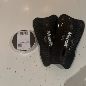 Monolit benskydd  - Säljer ett par svarta benskydd från Monolit, tillverkade i lätt kolfiber med snygg glansig finish och ergonomisk form. Perfekta för fotboll och kommer med vit sock tape från Select (1,9 cm x 15 m) 