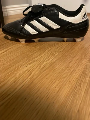 Svarta och vita adidas fotbollsskor - Säljer ett par klassiska adidas fotbollsskor i svart och vitt med de ikoniska tre ränderna på sidan. Skorna har snörning och platt sula med fasta dobbar för bra grepp på planen. Tillverkade i skinn för skön passform och känsla. Perfekta för dig som vill ha stil på fotbollsplanen.