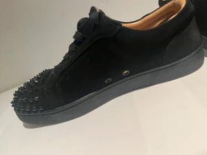Christian Louboutin svarta sneakers med nitar - Svarta sneakers från Christian Louboutin i mocka med coola svarta nitar på tån och klassisk röd sula. Skorna har rund tå, snörning och en platt sula. Insidan är beige och mjuk, perfekt för dig som vill sticka ut med en edgy vibe. Har dock ramlat en spik eller gått sönder. Skriv för mer bilder eller intresserad! Har inte boxen till den dock 