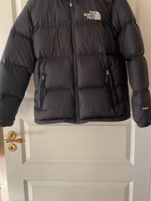Svart pufferjacka från The North Face - Säljer en svart pufferjacka från The North Face med 700-fill märkning. Jackan har två fickor med dragkedja, hög krage och logga på både fram- och baksidan. Perfekt för kalla dagar och har en clean, oversized look. OBS: barnstorlek