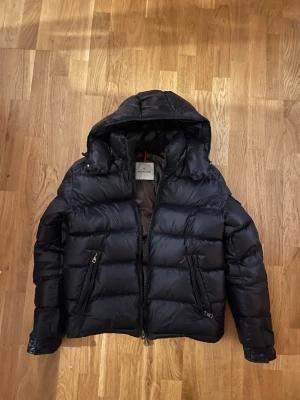 Navy blue Moncler pufferjacka med huva - Säljer en navy blue moncler Maya pufferjacka från Moncler med glansig finish och huva. Jackan har två framfickor med dragkedja, innerficka och Moncler-logga på ärmen. Insidan är brun/vinröd touch och har ett coolt serietryck. Perfekt för kalla dagar och riktigt snygg stil.