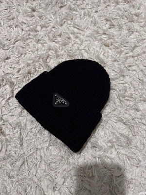 Svart ribbad mössa från Prada - Snygg svart mössa från Prada med ribbad stickning och ikonisk metalltriangel framtill. Mössan har en klassisk form med uppvikt kant och är tillverkad i mjukt stickat material som håller dig varm och stilren.