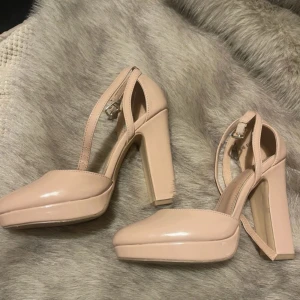 Beiga pumps med platå från New Look - Snygga pumps i beige/rosa lack från New Look med rund tå, hög blockklack och platåsula. Skorna har smala remmar med spänne runt ankeln för extra support. Perfekta för dig som vill ha både höjd och stabilitet. Materialet är glansigt syntet. 