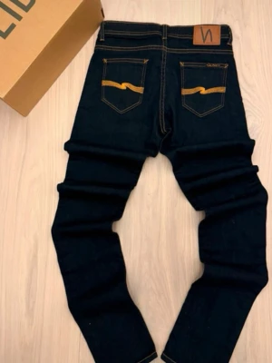 Mörkblå Nudie Jeans Skinny PRIS KAN DISKUTERAS ÖPPEN FÖR MÅNGA BILLIGA PRIS! - Säljer ett par mörkblå skinny jeans från Nudie Jeans med klassisk orange söm och broderad logga på bakfickorna. Jeansen har normal passform, låg midja och är tillverkade i slitstarkt bomullsmaterial. Perfekt för dig som gillar stilrena och tajta jeans. PRIS KAN DISKUTERAS!