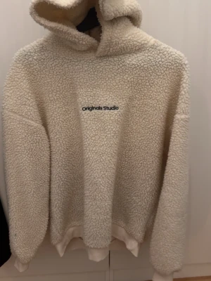 Beige teddyhoodie från Jack & Jones - Mysig teddyhoddie i beige från Jack & Jones Originals. Lång ärm och broderad text på bröstet. Perfekt för kalla dagar när du vill vara både varm och snygg. 