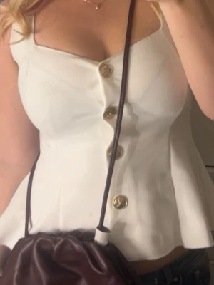 Vit linnetopp med guldfärgade knappar - Snygg vit linnetopp med breda axelband och guldfärgade knappar framtill. Toppen har en lätt utsvängd form och markerad midja, vilket ger en feminin siluett. Perfekt att styla med jeans eller kjol för en trendig look.