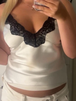 Vit satin linnetopp med spets - Säljer en snygg vit linnetopp i satin med svart spetsdetalj längs v-ringningen. Toppen är ärmlös och har en croppad passform som ger en trendig look. Perfekt att matcha med jeans eller kjol för en cool stil.