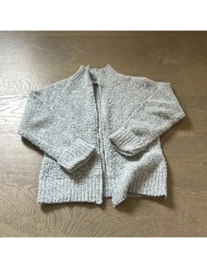 Grå ullmix stickad kofta från Fransa - Mysig grå ullmixad kofta från Fransa med grovstickad struktur och ribbade muddar. Koftan har lång ärm och är öppen framtill utan knappar eller dragkedja. Perfekt att slänga på sig när det är lite kyligt ute.