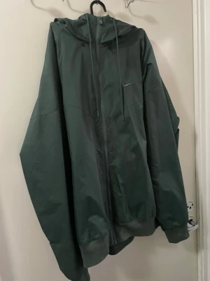Grön Nike vindjacka med huva - Säljer en grön Nike vindjacka i loose fit med huva och dragkedja framtill. Jackan har ribbade muddar vid ärmslut och nederkant, samt snörning vid huvan. Materialet är lätt och syntetiskt, perfekt för blåsiga dagar. Nike-logga broderad på bröstet.